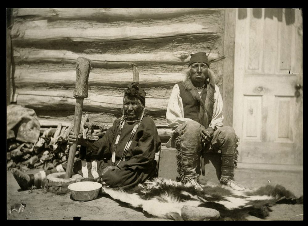 Yellow Nose sits with her husband Bear on the Water in front of their home while she uses a mortar to pound dried corn into flour.