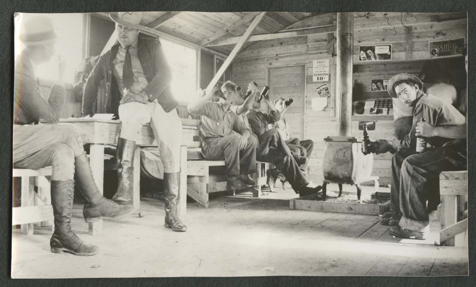 Men in building with stove drinking something from a bottle and talking with two men with hats and high boots, site unknown