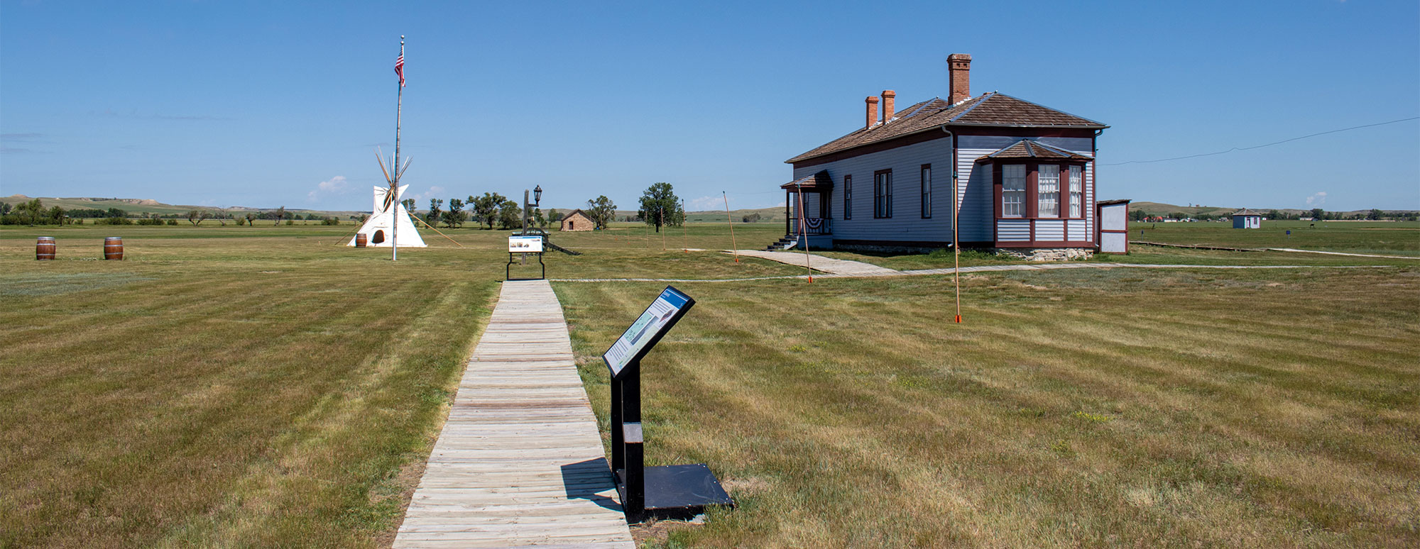 A wooden walkway with interpretive signage on the side leads to an old, gray building with red trim. There is also a tipi in the background on the other side.