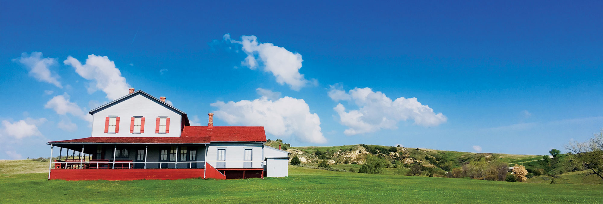 light gray house with a red porch and roof sits on green grass with a blue sky and buttes in the background