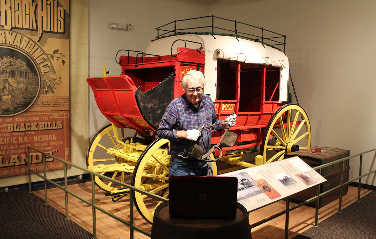 A man stand holding some sort of old belt. Behind him is a red, wooden carriage with yellow wheels.